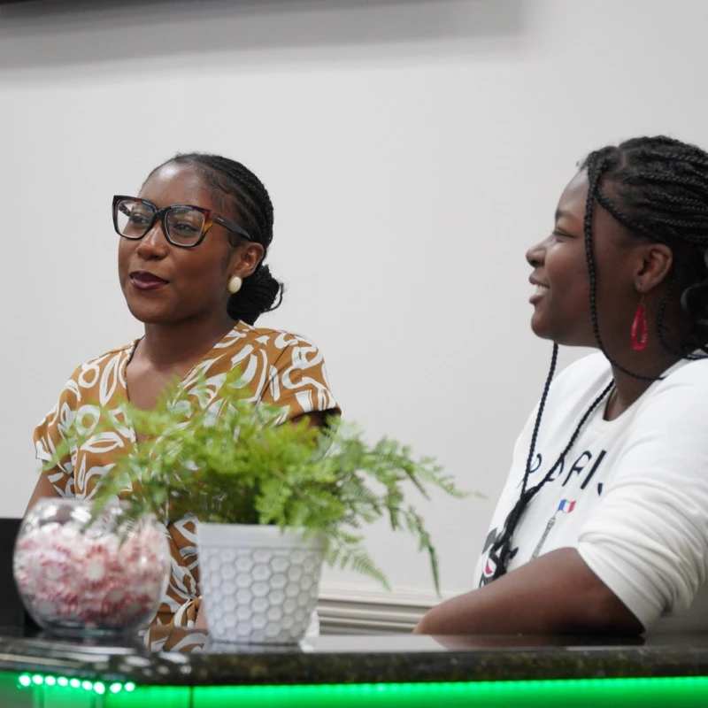 Two Black women are sitting at a desk with a plant on it. One woman wears glasses and a patterned shirt, and the other wears a white shirt and is smiling.